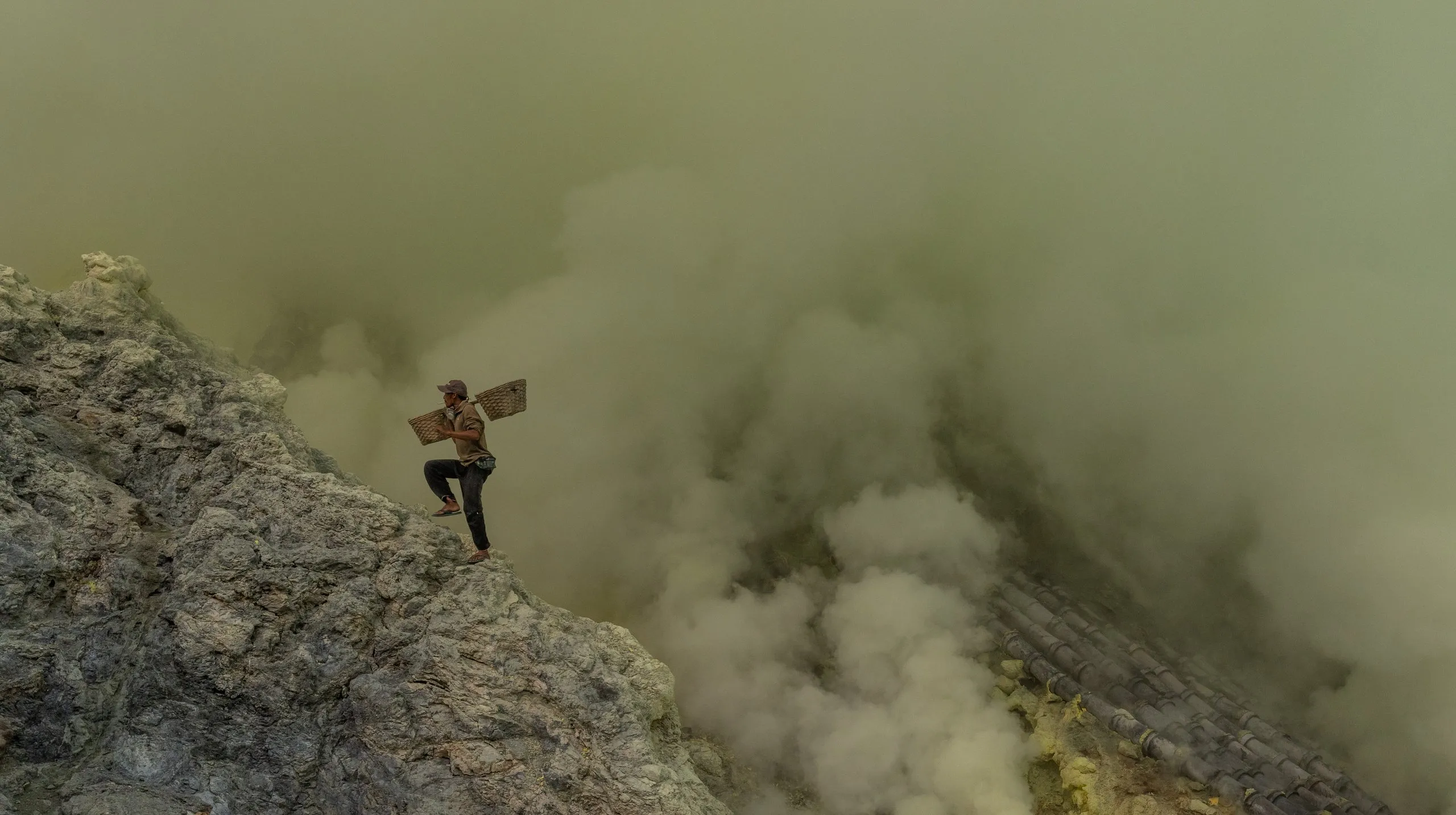 Sulfur miner climbing the steep crater wall of Kawah Ijen with loaded baskets, smoke rising behind him